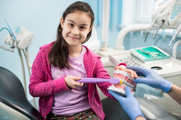 girl in dentist chair showing proper tooth-brushing using dental jaw model and big toothbrush